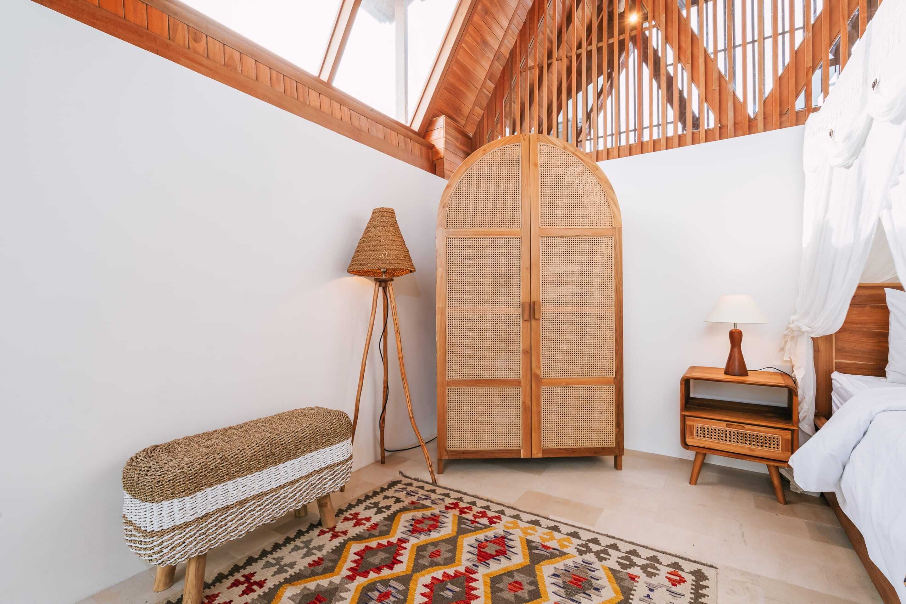 A cozy bedroom with a wooden ceiling, a woven bench, a rattan wardrobe, a patterned rug, and a bedside table with a lamp.