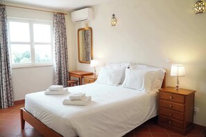 A neatly made bed with white linens, a bedside table with a lamp, a mirror, and a window with curtains.