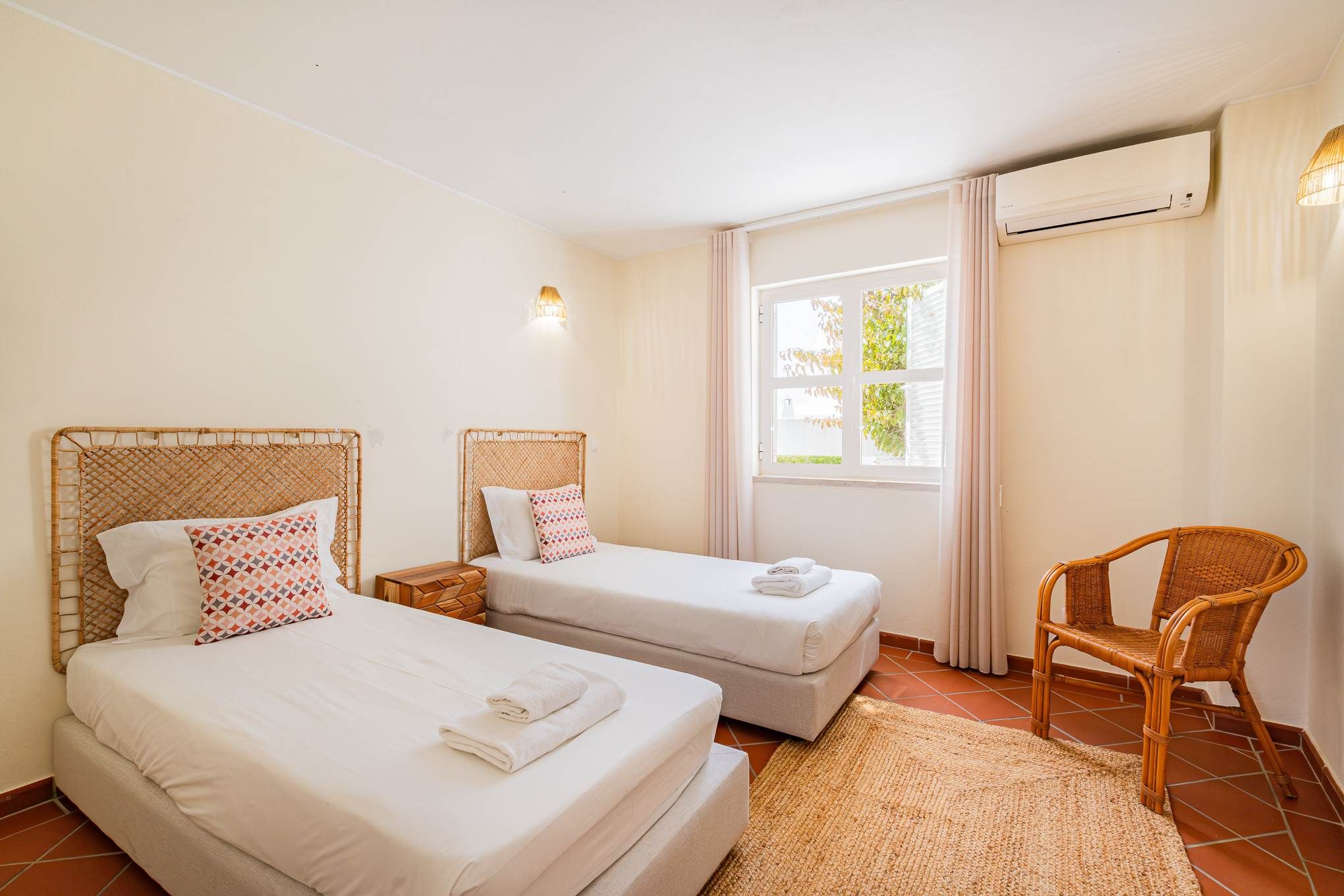 Quarto com duas camas de solteiro, uma cadeira, janela com cortinas e ar condicionado.