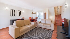 Un salon avec un canapé, une table basse et un téléviseur. On y trouve également un coin repas avec des chaises et une table. Une cuisine est visible au fond.
