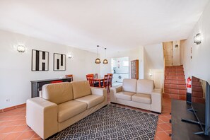 Un salon avec un canapé, une table basse et un téléviseur. On y trouve également un coin repas avec des chaises et une table. Une cuisine est visible au fond.