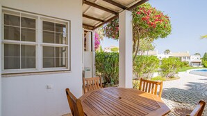 A covered patio with a wooden table and chairs, a window with blinds, and a view of a swimming pool and greenery. - Villa Lychee - 350 m from the beach (Guia)