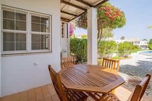 Une terrasse couverte avec une table et des chaises en bois, une fenêtre avec des stores et une vue sur une piscine et la verdure.
