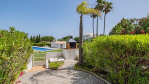 Un chemin pavé mène à une piscine, où se trouvent des chaises longues, un espace barbecue et une végétation luxuriante.