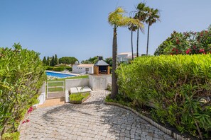 A paved pathway leading to a swimming pool with lounge chairs, a barbecue area, and lush greenery.