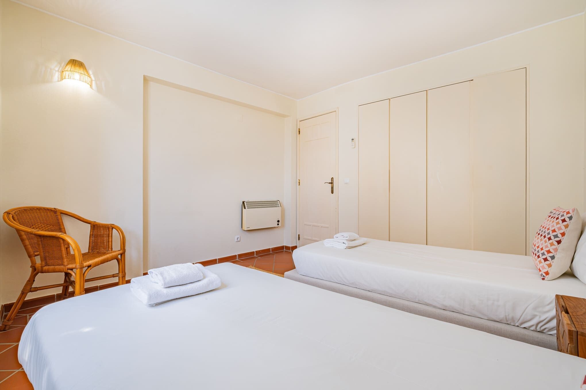 A hotel room with two beds, a chair, a heater, and a wall-mounted light fixture.