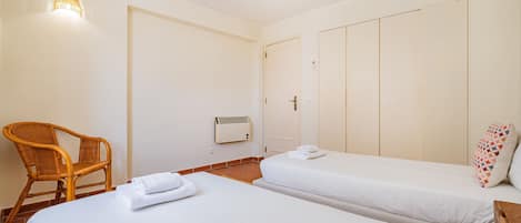 A hotel room with two beds, a chair, a heater, and a wall-mounted light fixture.