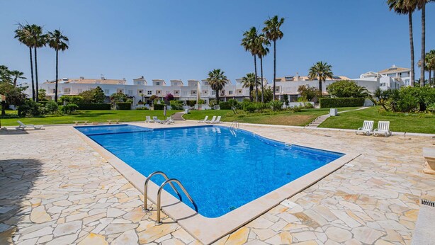 Une piscine dotée d’une terrasse en pierre, entourée de palmiers et bordée d’une rangée de bâtiments blancs.