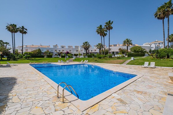 A swimming pool with a stone deck, surrounded by palm trees and a row of white buildings.
