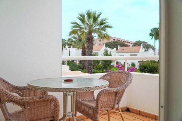 A balcony with wicker chairs and a glass table, overlooking a garden with palm trees and buildings.
