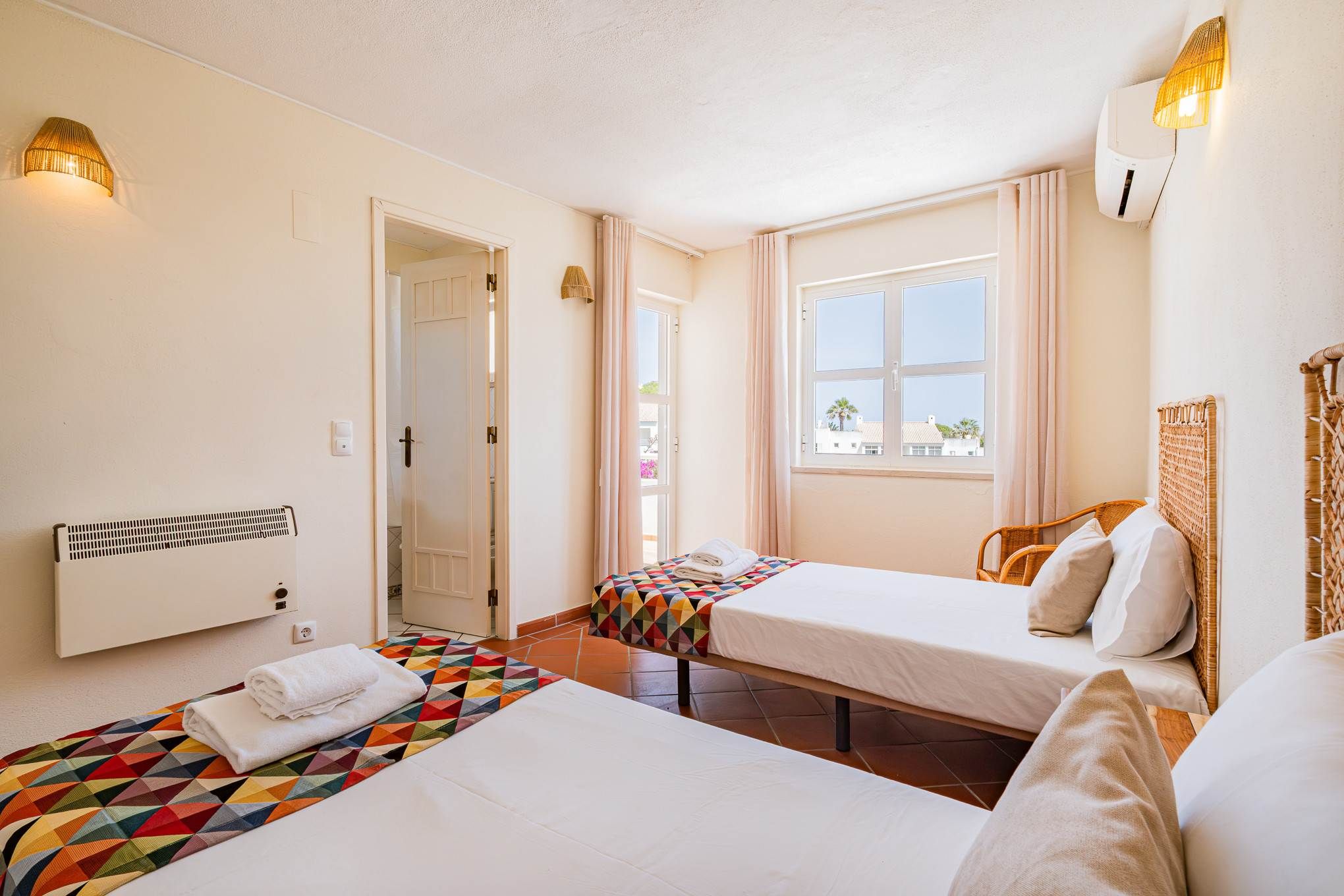 A hotel room with two beds, a window with curtains, a radiator, and a wall-mounted lamp.