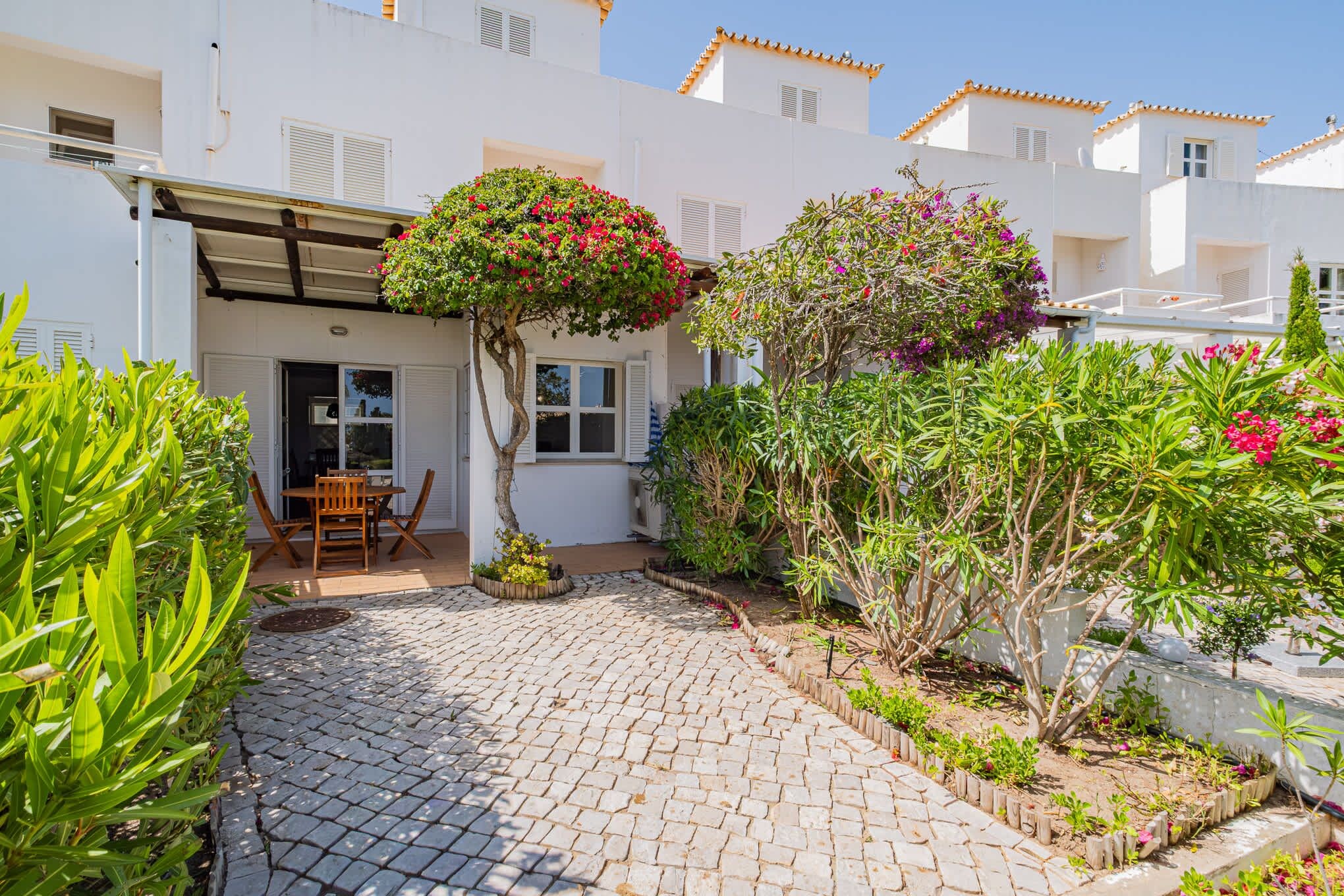 A courtyard with a cobblestone path, a wooden table and chairs, and various plants and shrubs.