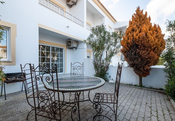 A courtyard with a round metal table and chairs, a bench, and a tall tree.