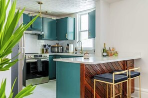 Una cocina con armarios color turquesa, una isla central con taburetes, electrodomésticos de acero inoxidable y un ventanal que proporciona luz natural.
