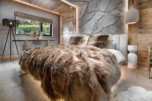 A modern bedroom with a large bed, a wooden ceiling, and a view of greenery outside.