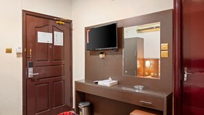 Een hotelkamer met een houten deur, een flatscreen televisie aan de muur, een spiegel met aanrichtplaats, een kleine prullenbak en een bankje.