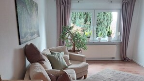 Een woonkamer met een bank, een houten bijzettafeltje, een groot raam met gordijnen, en een potplant.