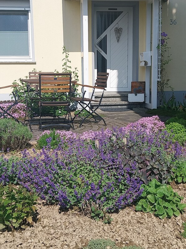 A patio with a wooden bench and chairs, surrounded by a garden with purple flowers.