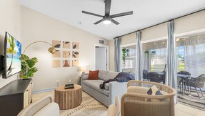 Un salon moderne avec un canapé, une table basse et des fauteuils en osier. Un ventilateur de plafond et un téléviseur sont fixés au mur.