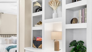 Une pièce bien rangée, avec un lit, une étagère blanche ornée d’objets décoratifs et une plante en pot.
