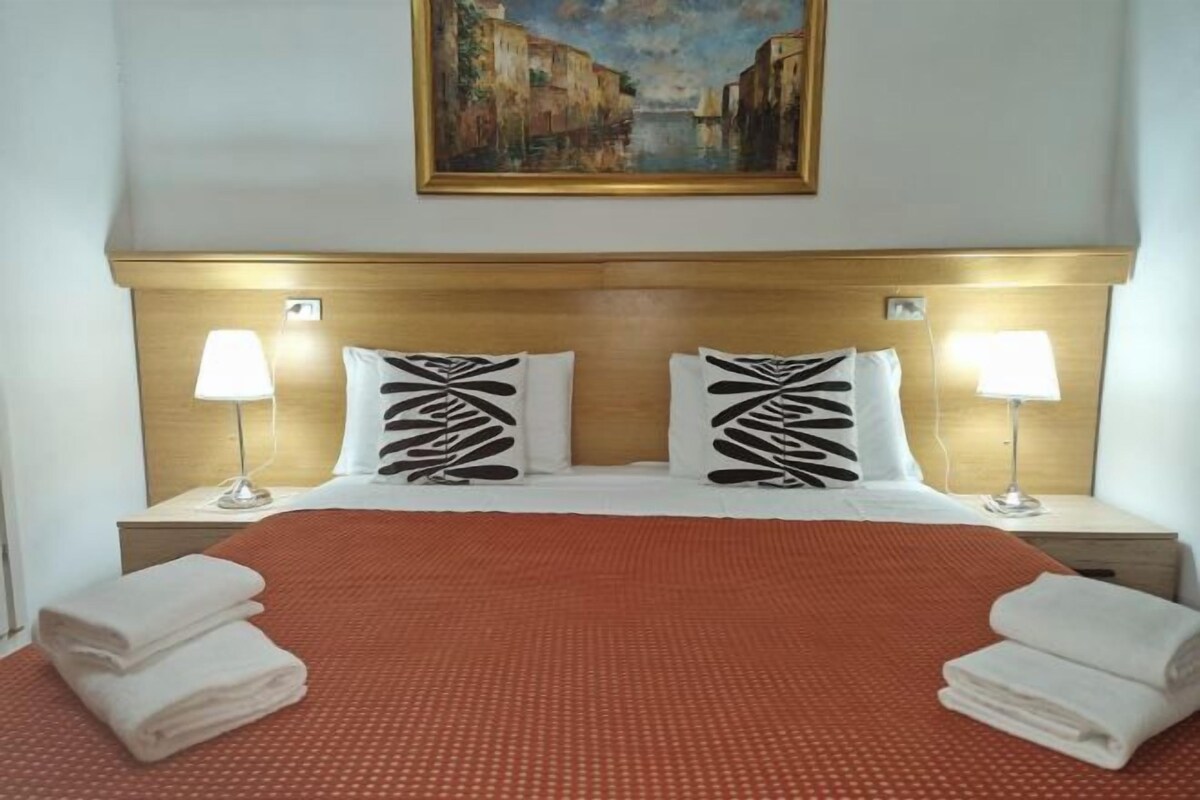 A hotel room with a large bed, two bedside lamps, a painting above the bed, and neatly folded towels on the bed.