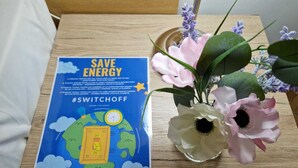 A poster on a wooden table promoting energy conservation with a globe illustration and a clock, alongside a vase with flowers. - Hotel Tito (Mestre)