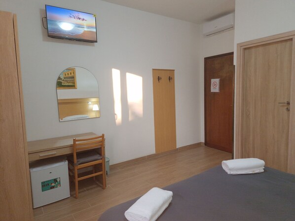 A hotel room with a bed, a desk with a chair, a small fridge, a television mounted on the wall, and a door with a sign.