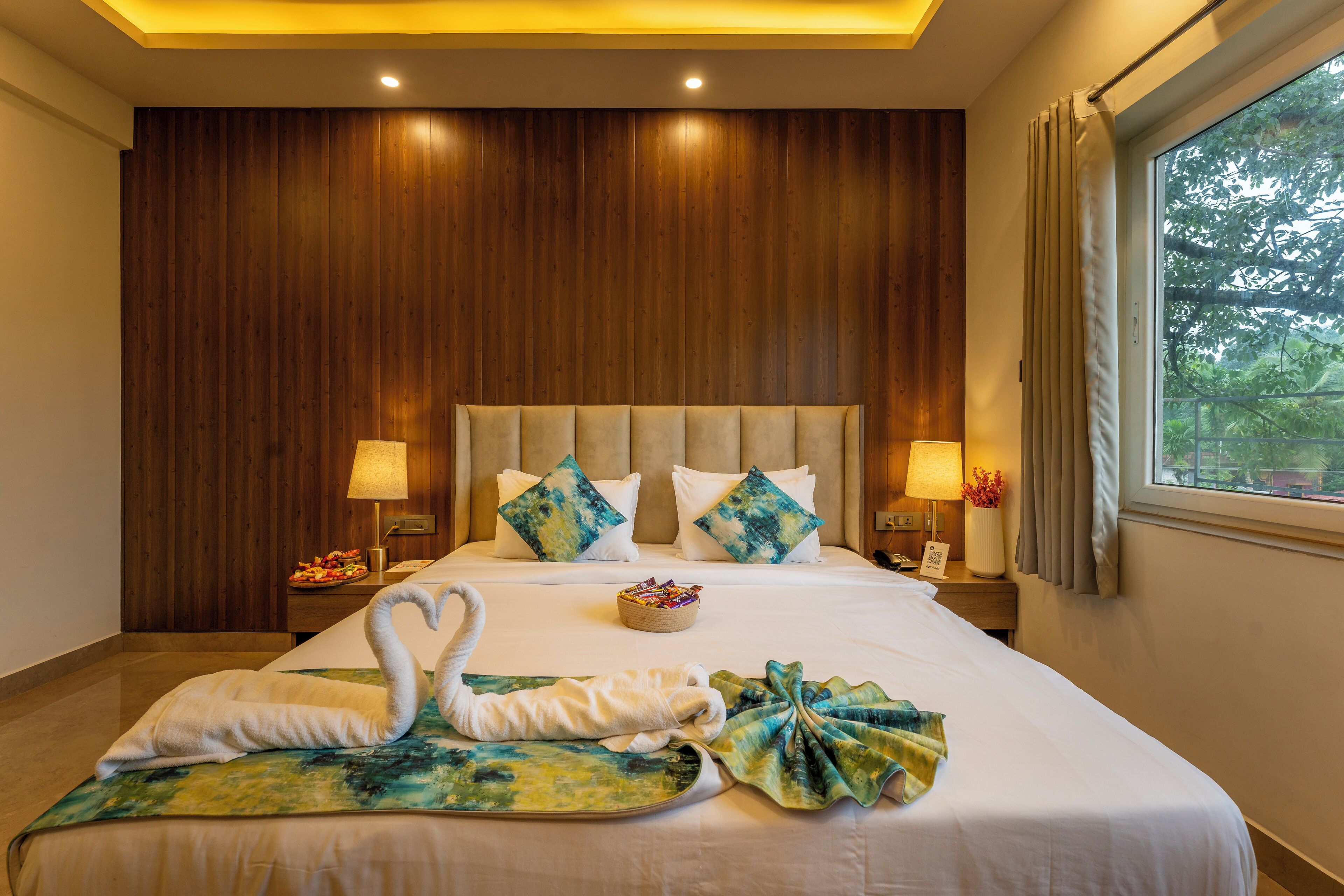 Habitación de hotel con una cama grande, paneles de madera en las paredes, dos lámparas de noche, una mesita con un frutero y una ventana con cortinas.