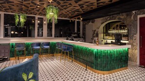 A bar area with a green illuminated bar counter, blue bar stools, and hanging greenery. - The Eden Inn By The Saga Stays (Anjuna)