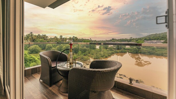 A balcony with wicker furniture, a glass table, and a view of a river and greenery. - The Eden Inn By The Saga Stays (Anjuna)