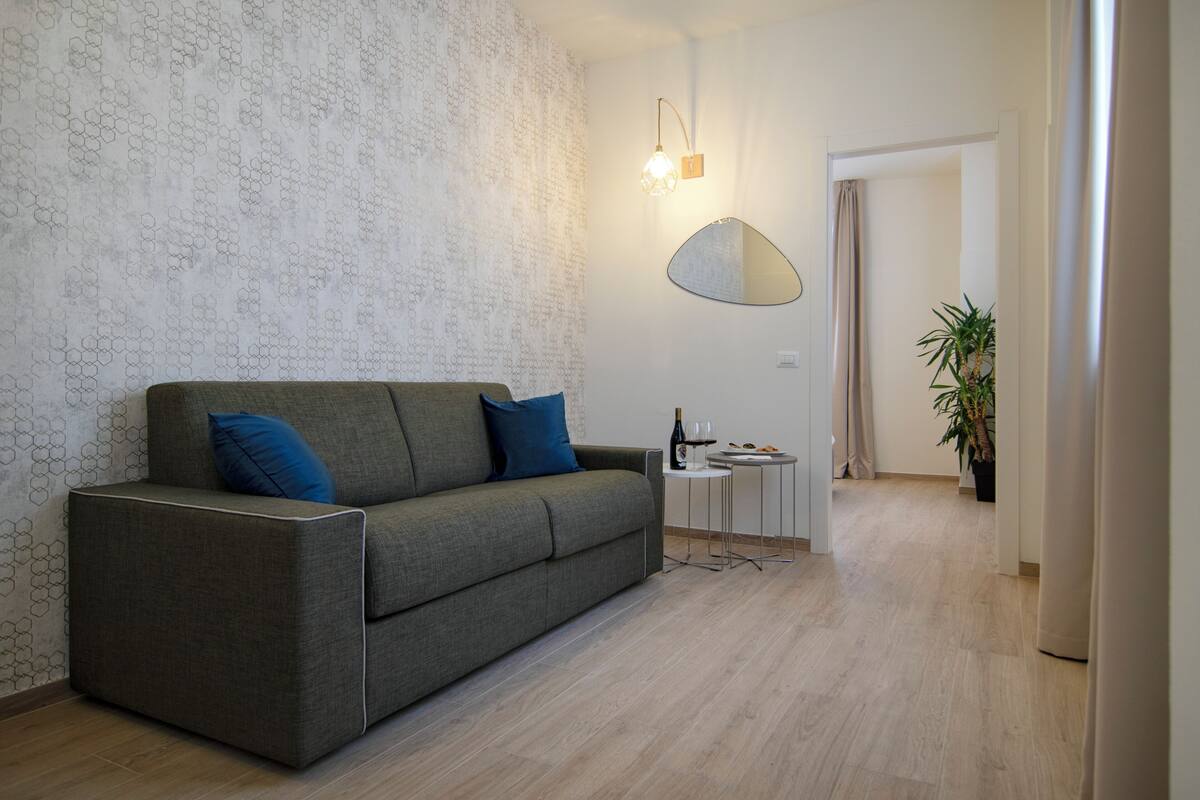 Ein modernes Wohnzimmer mit einem grauen Sofa, einem kleinen Beistelltisch, einem Spiegel und einer Topfpflanze.