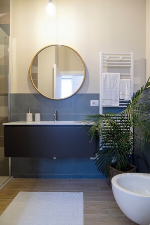 A modern bathroom with a dark blue vanity, a round mirror, a white towel rack, and a large white basin. - GIGI SUITES&APARTMENTS (Stresa)