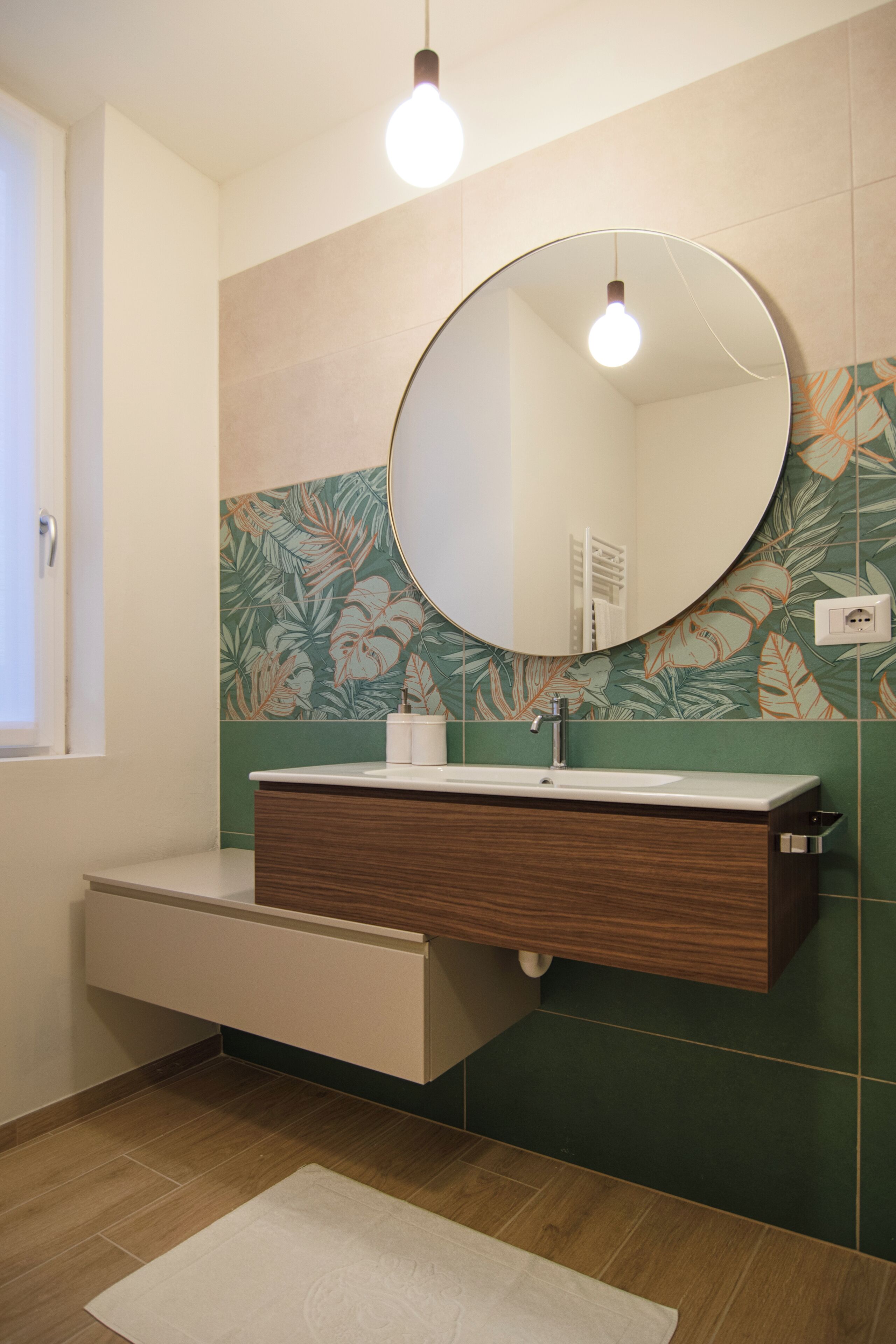 A bathroom with a wooden vanity, a round mirror, and a green floral tile backsplash.
