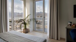 Quarto de hotel com cama, planta em vaso, vista de montanhas e lago através de janelas amplas, e uma pequena mesa com uma tigela.