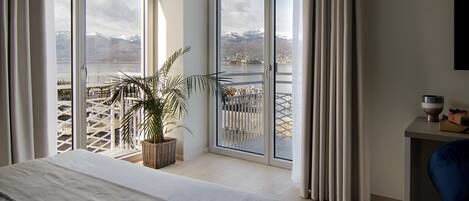 A hotel room with a bed, a potted plant, a view of mountains and a lake through large windows, and a small table with a bowl.