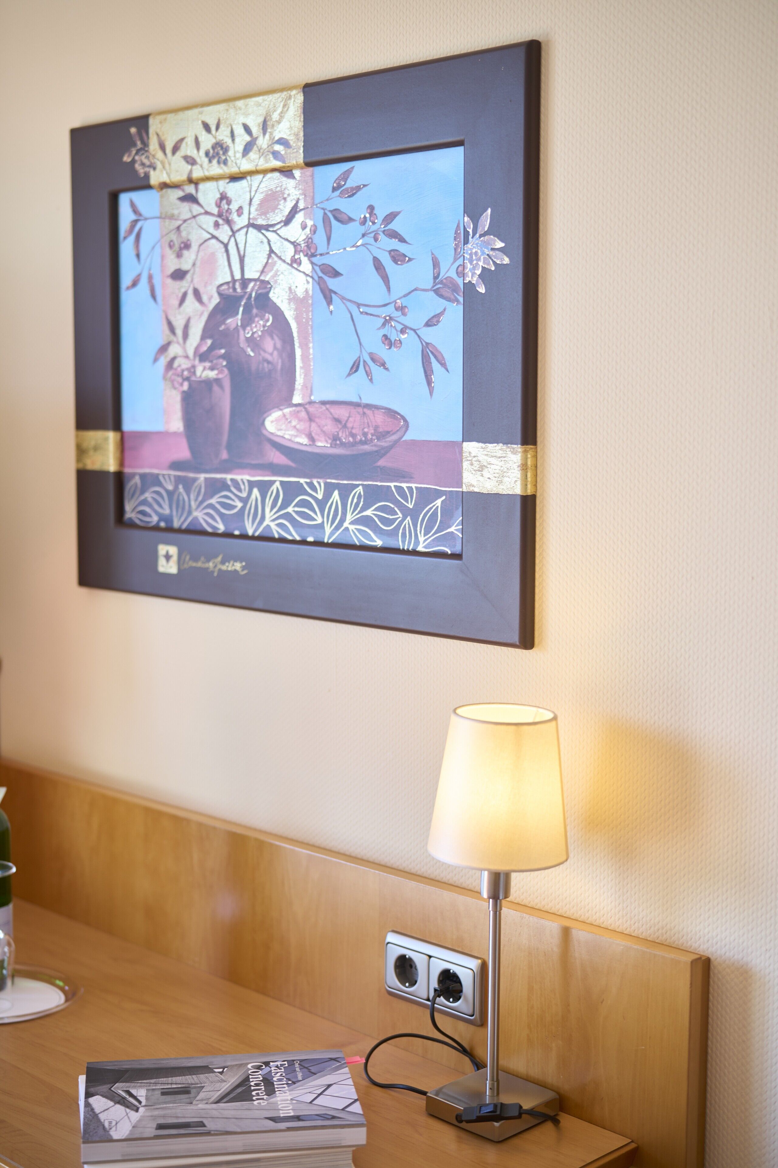 A framed artwork of a vase and branches on a wall, a bedside table with a lamp, and a book.