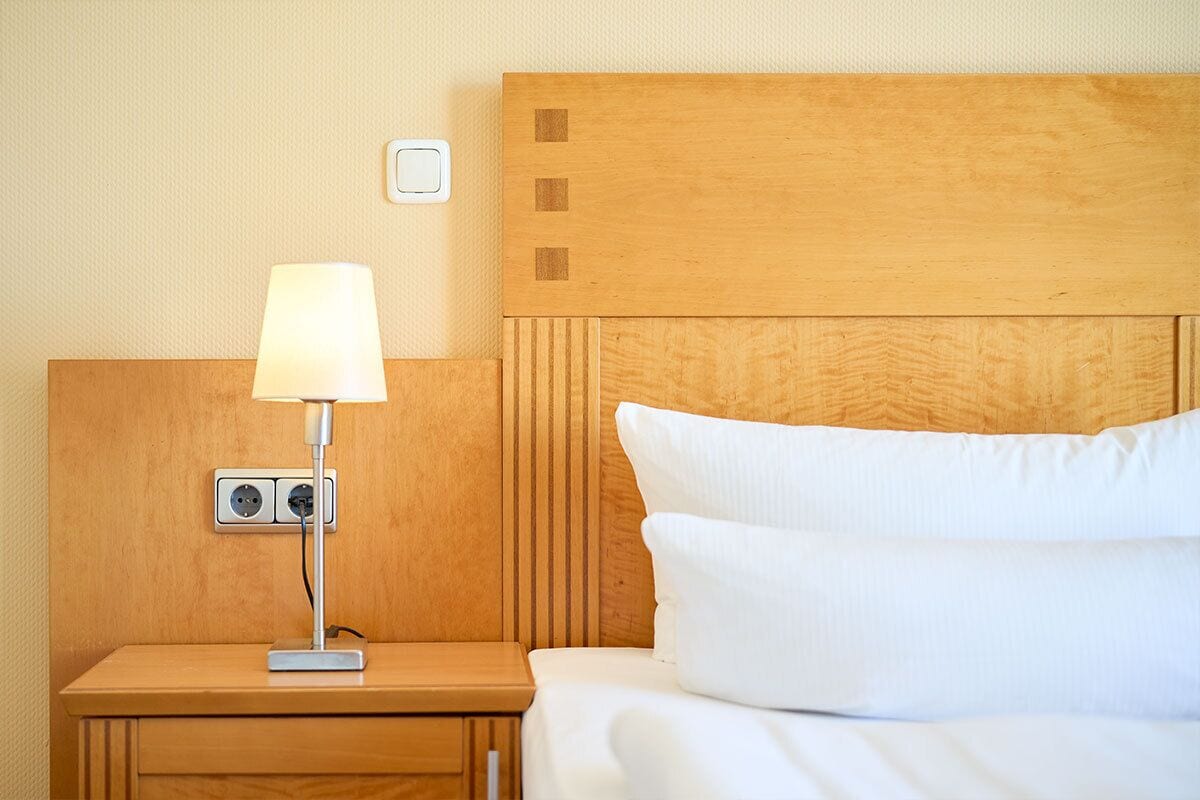 A neatly made bed with white linens, a wooden nightstand with a lamp, and a wall socket.