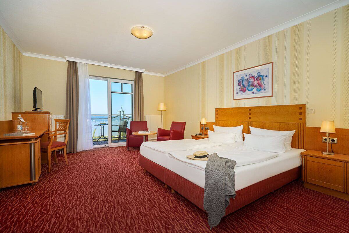 A hotel room with a large bed, a desk, a chair, a small table, and a balcony with a view.