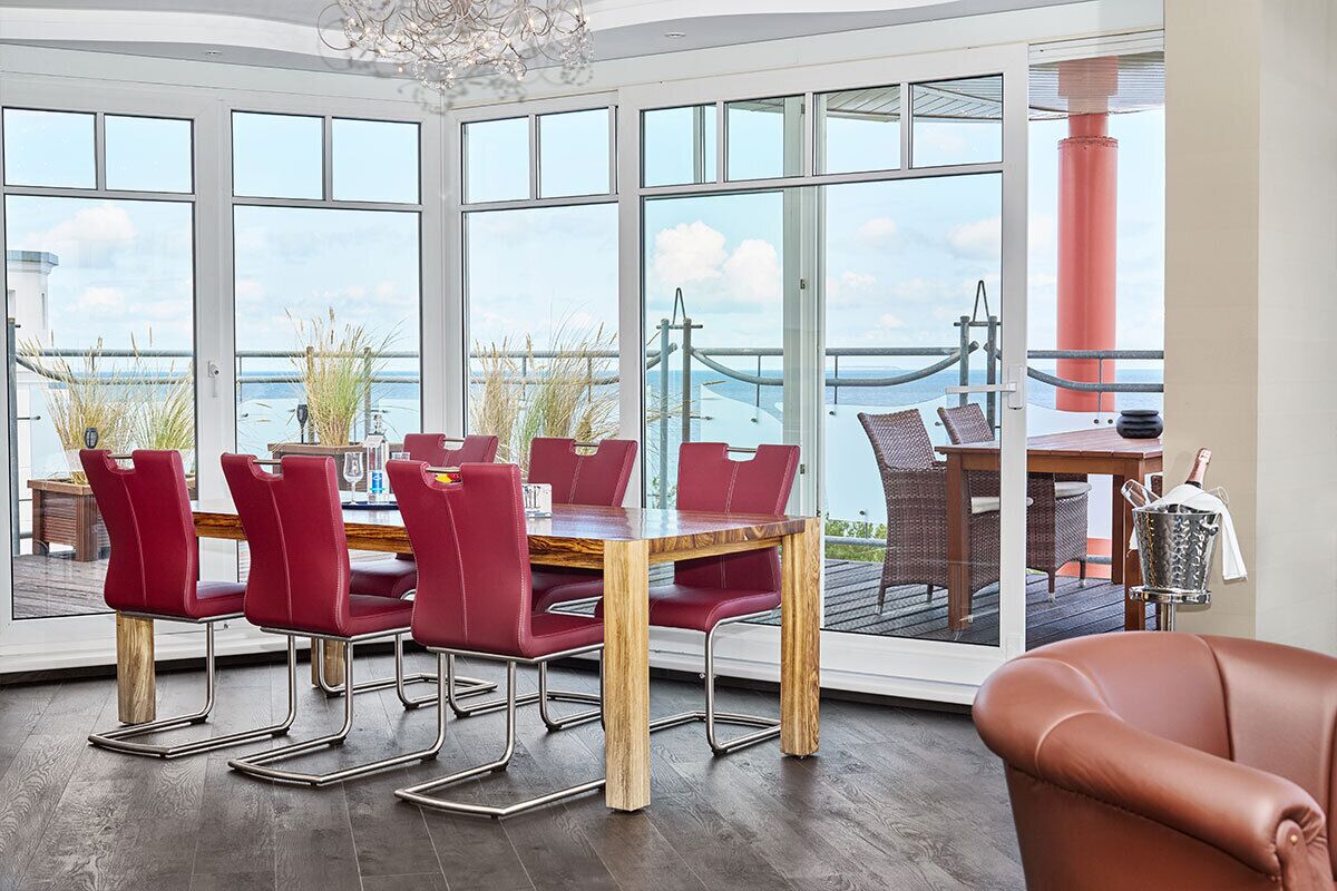 A dining area with a large table and red chairs, large windows with a view of the sea and a bridge, and a wooden floor.
