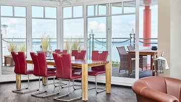 A dining area with a large table and red chairs, large windows with a view of the sea and a bridge, and a wooden floor.