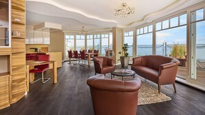 A spacious living area with a large window overlooking a body of water, a wooden dining table with red chairs, and leather sofas. - Strandhotel Prinz von Preussen (Zinnowitz)