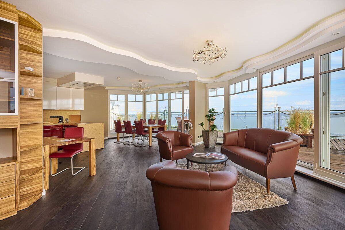 A spacious living area with a large window overlooking a body of water, a wooden dining table with red chairs, and leather sofas.