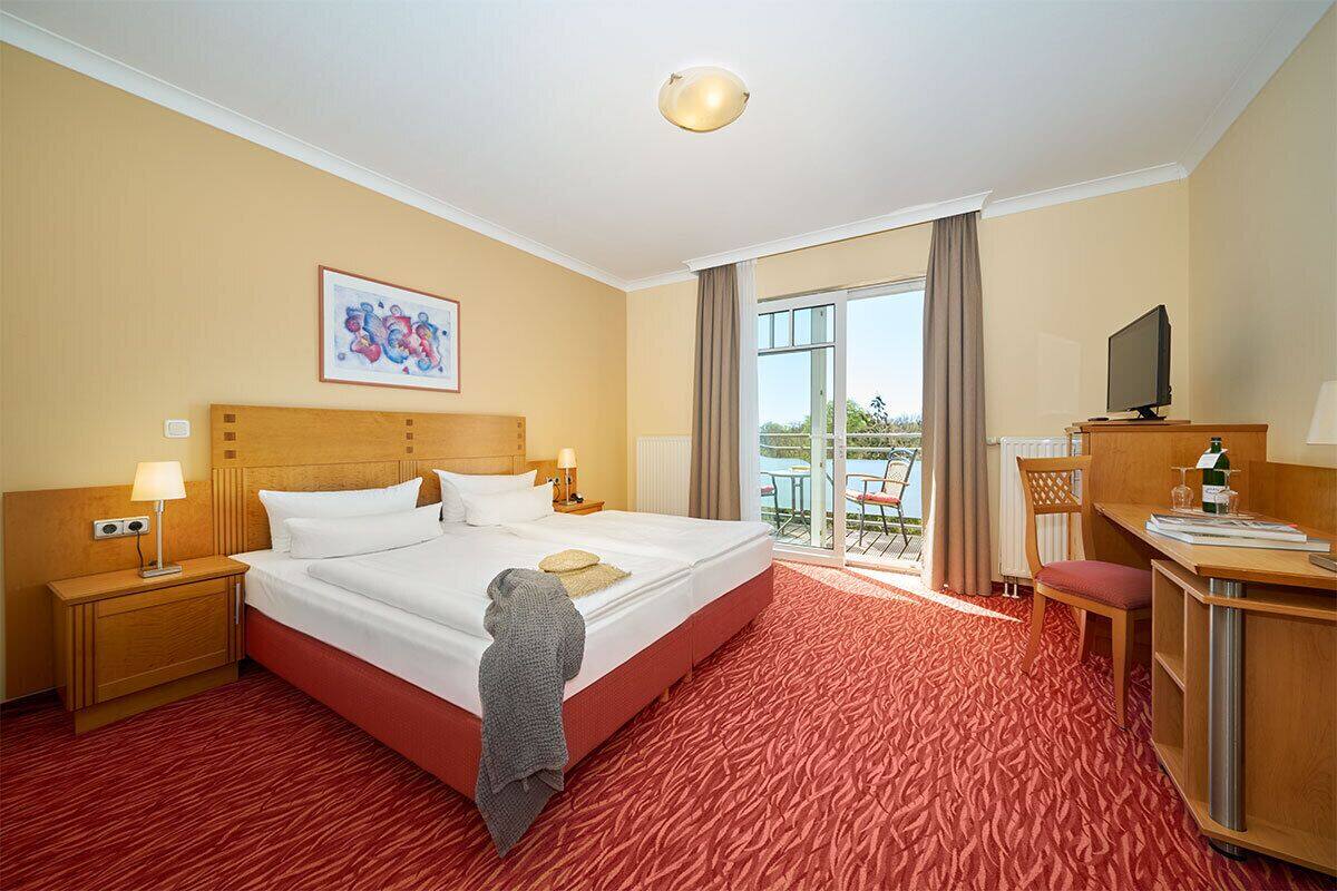 A hotel room with a large bed, a desk with a chair, a television, and a balcony with a view.