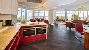 A modern kitchen with a red island, built-in ovens, and a large window overlooking the sea. - Strandhotel Prinz von Preussen (Zinnowitz)