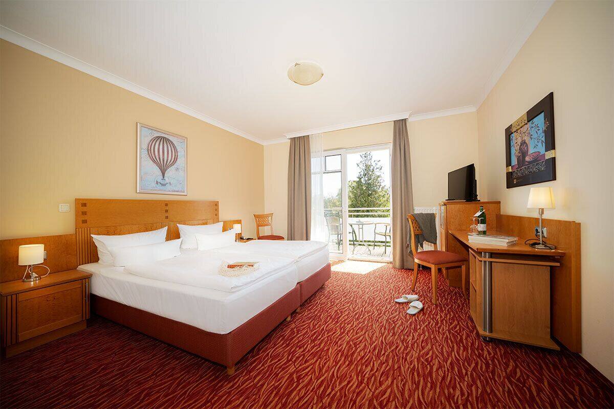 A hotel room with a large bed, a desk, a television, and a balcony with a view of trees.