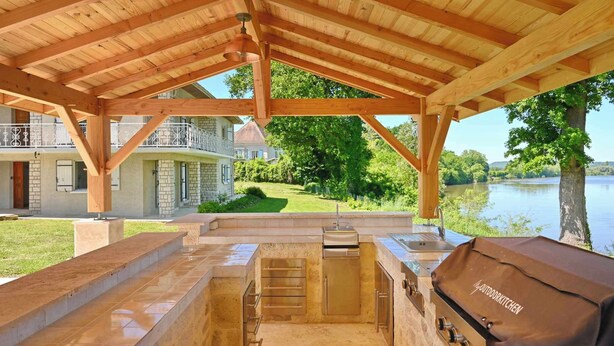 An outdoor kitchen with a grill, built-in sink, and a view of a lake and house in the background.