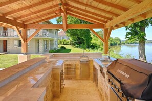 Une cuisine extérieure équipée d’un barbecue, d’un évier intégré et offrant une vue sur un lac et une maison en arrière-plan.