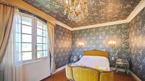 A bedroom with a floral wallpaper, a wooden floor, a yellow bed, a chandelier, and a window with curtains.