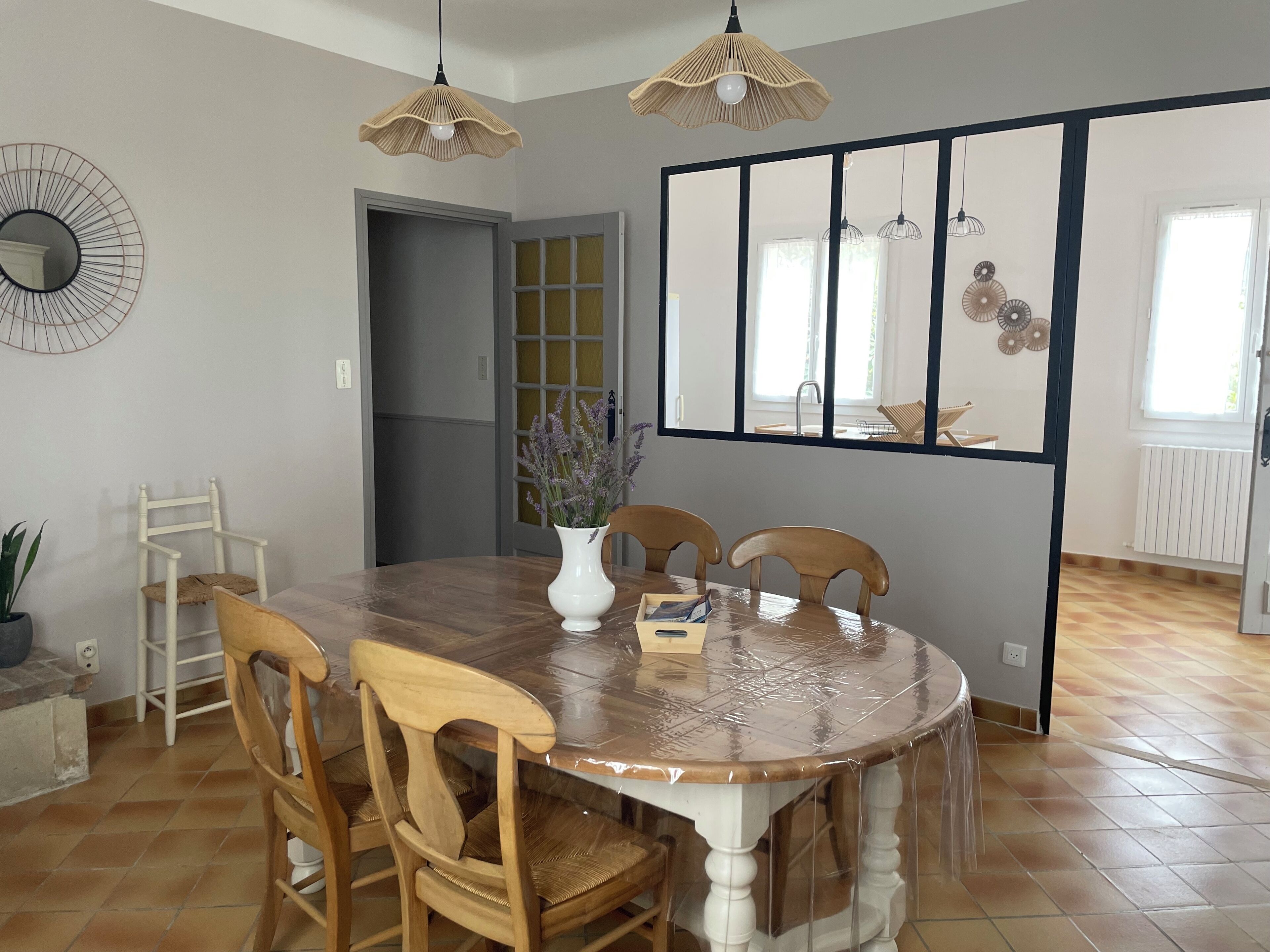 A dining area with a round wooden table, four wooden chairs, a white vase with flowers, and two hanging wicker lamps.
