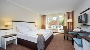 A hotel room with a large bed, a desk with a chair, a TV, and a window with a view of the outside. - Strandhotel Luise von Preussen (Zinnowitz)